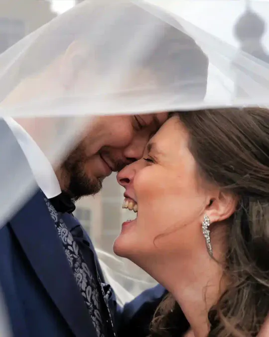 Elke en Thibaut lachen, voorhoofd tegen voorhoofd, onder de sluier van de bruid