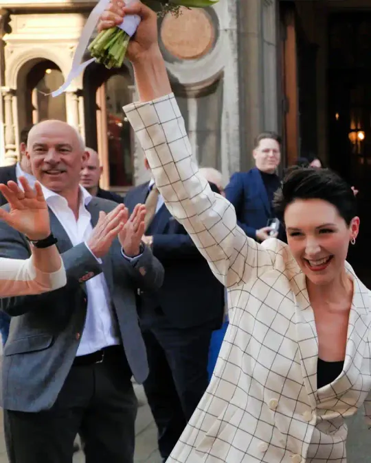 Elita steekt juichend haar boeket omhoog, omringd door applaudisserende gasten